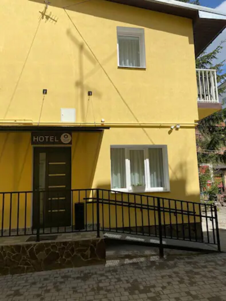 The exterior of a hotel is shown, featuring a bright yellow facade. A modern entrance is highlighted by a metal railing leading up to a door. Two windows are visible, allowing natural light to enter. The surrounding area consists of cobblestone paving.