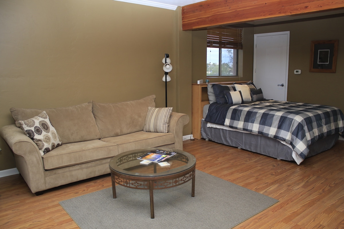A spacious guest suite features a comfortable sofa and a glass-top coffee table on a patterned area rug. A queen-sized bed is dressed in a plaid duvet and is positioned beside a window with wooden blinds. A simple, neutral tone decor creates a relaxing ambiance.