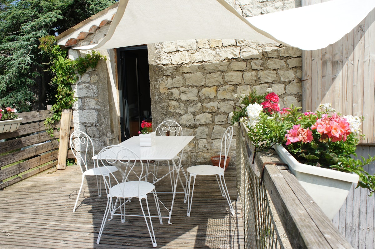 A shaded terrace features a white dining table surrounded by four matching chairs. Flower boxes with colorful blooms accent the wooden railing, creating a bright and inviting outdoor space. The stone walls of the building provide a rustic backdrop.