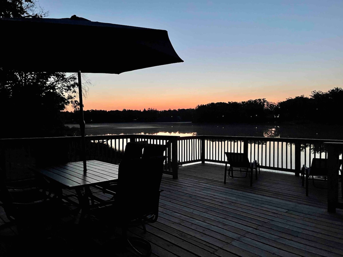 A spacious deck is shown overlooking a serene lake at dusk. Silhouettes of chairs and a table are visible, with an umbrella providing shade. The horizon features soft hues of orange and purple as the sun sets behind the trees.