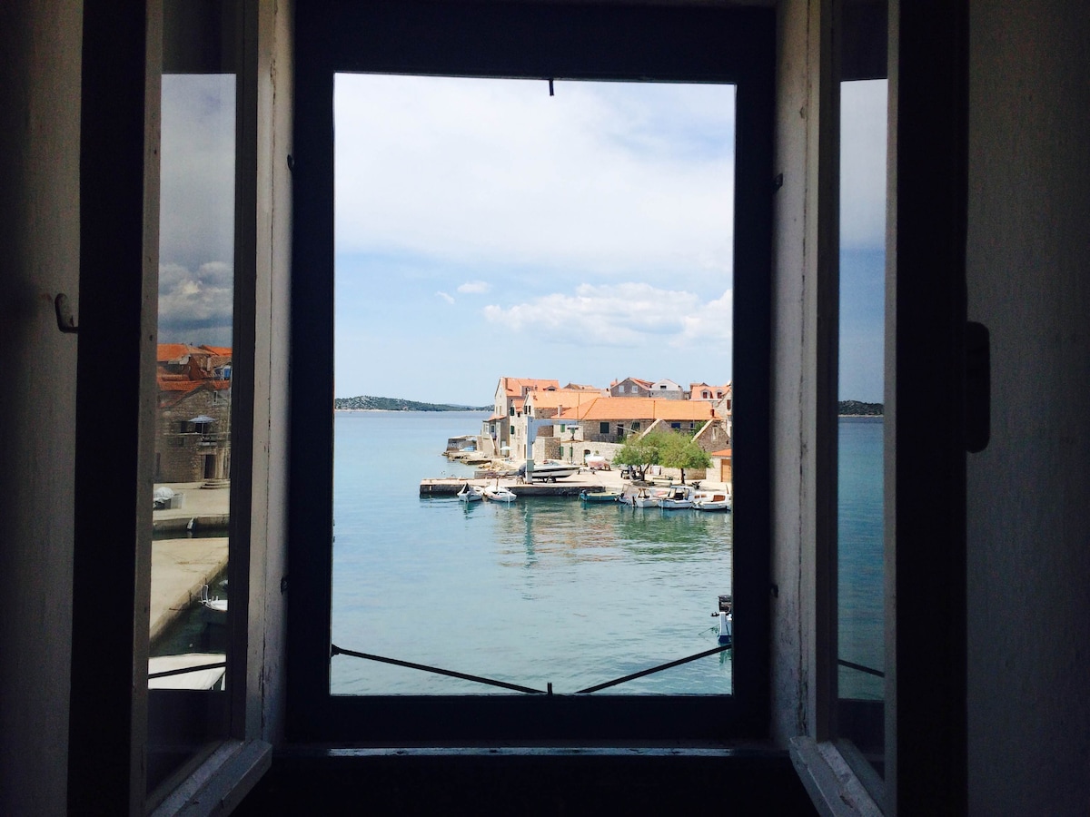 A view through an open window displays a serene coastal scene, where calm waters reflect the surrounding buildings and boats. The distant shoreline is dotted with charming structures, framed by the window's rustic wooden panes.