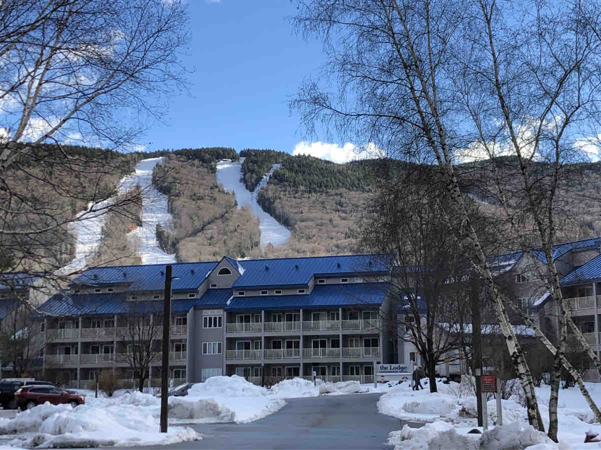 The image displays the Lodge at Lincoln Station, featuring a mountainous backdrop with ski trails visible on the slopes. A clear blue sky complements the scene, and snow is present in the surrounding area, suggesting a winter setting.
