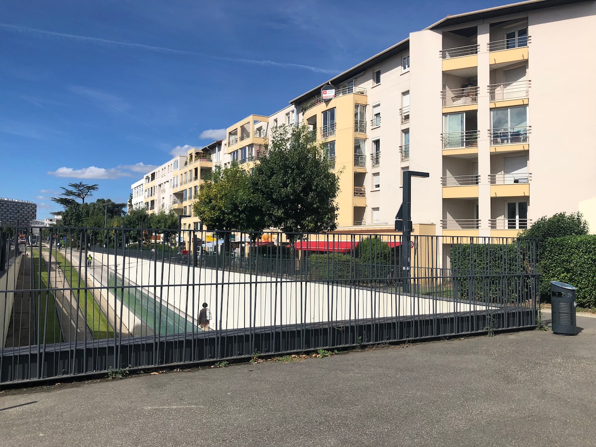 A view of a modern apartment complex featuring light-colored façades and multiple balconies. A gated area is visible in the foreground, adjacent to a wide green space. Lush trees are planted alongside the pathway, enhancing the residential setting.