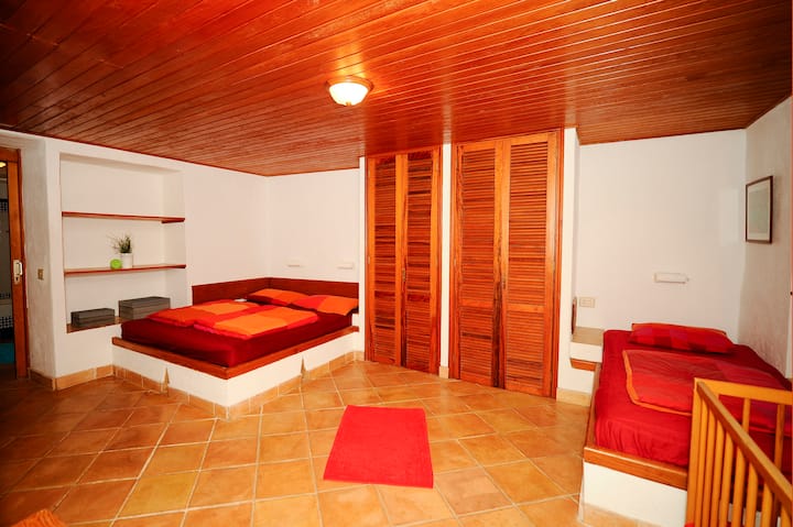 Голяма спалня на приземния етаж на сградата с двойно легло и единично легло, до която се стига от горния етаж до стълбището на открито или от терасата. Снимка, направена от входа.