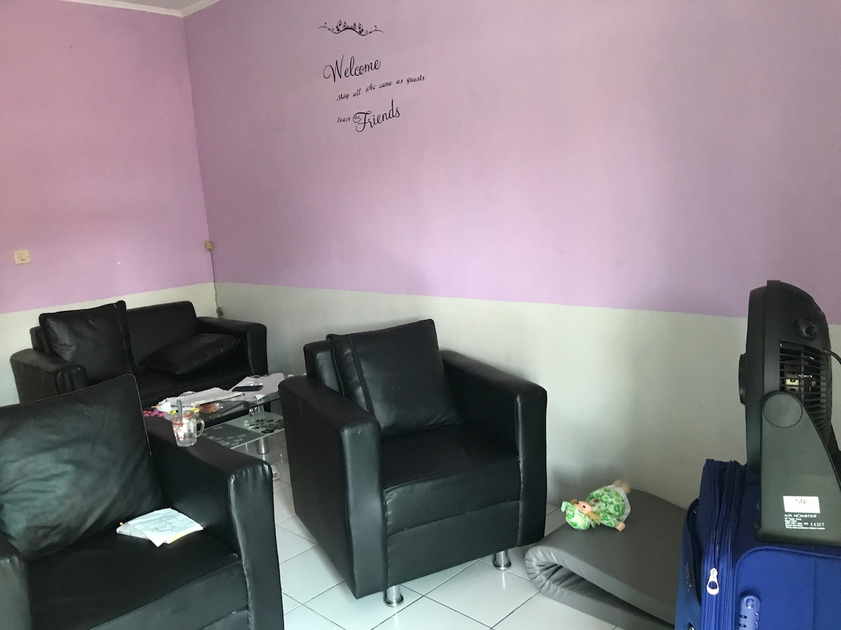 A cozy living area features two black armchairs and a glass coffee table, with a purple wall creating a soothing backdrop. A suitcase sits nearby, and a fan is positioned on the floor, facilitating airflow in the space. Relaxed and functional arrangements are evident.