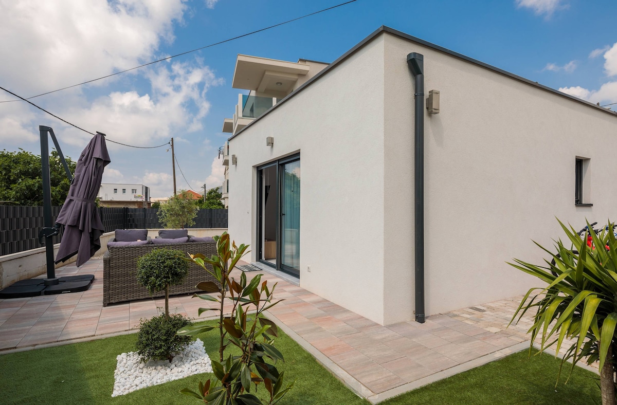A modern exterior of the apartment is showcased, featuring a spacious patio area with outdoor seating. Lush greenery, including a small shrub and potted plants, adorns the landscaped yard. The building's clean lines and neutral color palette reflect contemporary design elements.