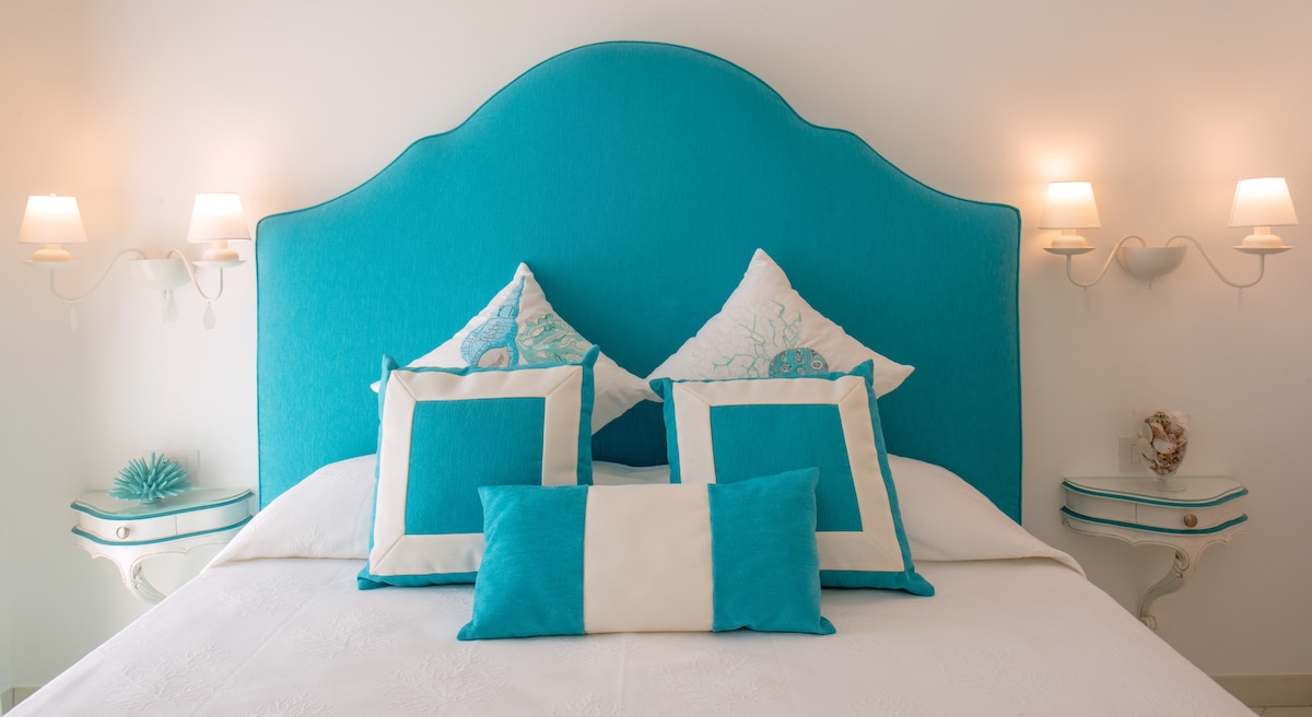 A spacious bed is adorned with turquoise and white decorative pillows. The headboard features a soft, curved design in a vibrant turquoise hue, complemented by wall sconces on either side. Nightstands with intricate detailing provide functional accents.