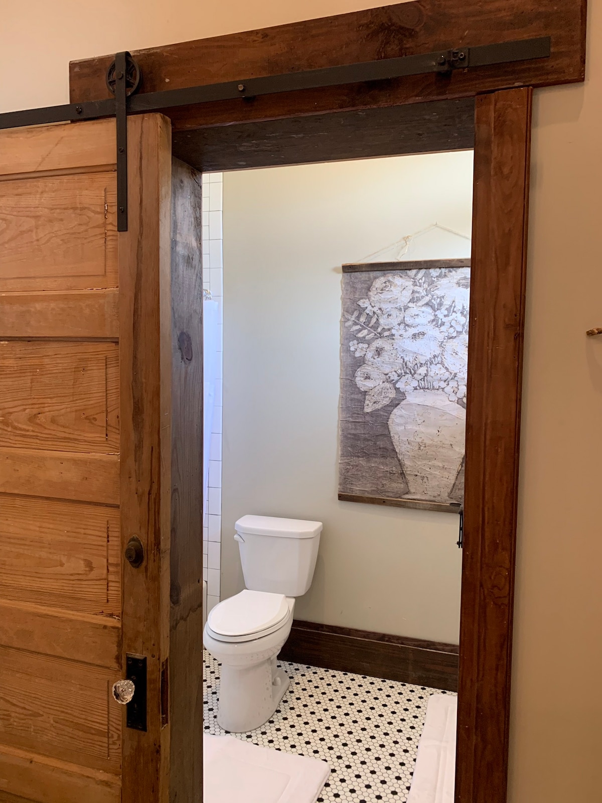 A view of a bathroom framed by a rustic sliding barn door. The space features a white toilet and a decorative wall hanging depicting a vase of flowers. Black and white patterned floor tiles add a stylish touch to the room.