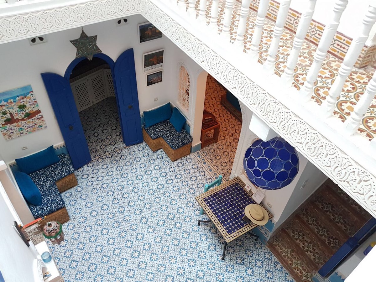 An open-air patio features intricate blue tile patterns under a bright ceiling. Comfortable seating areas are arranged near the walls, with a traditional table in the center. Decorative artwork is displayed throughout, and a staircase leads to the upper levels.