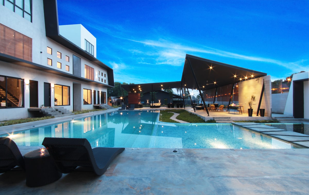 An outdoor area features a modern swimming pool with various lounging options nearby. A spacious shaded seating area is visible in the background, complemented by illuminated landscape lighting. The evening sky presents soft blue hues, creating a relaxing atmosphere for gathering and leisure.