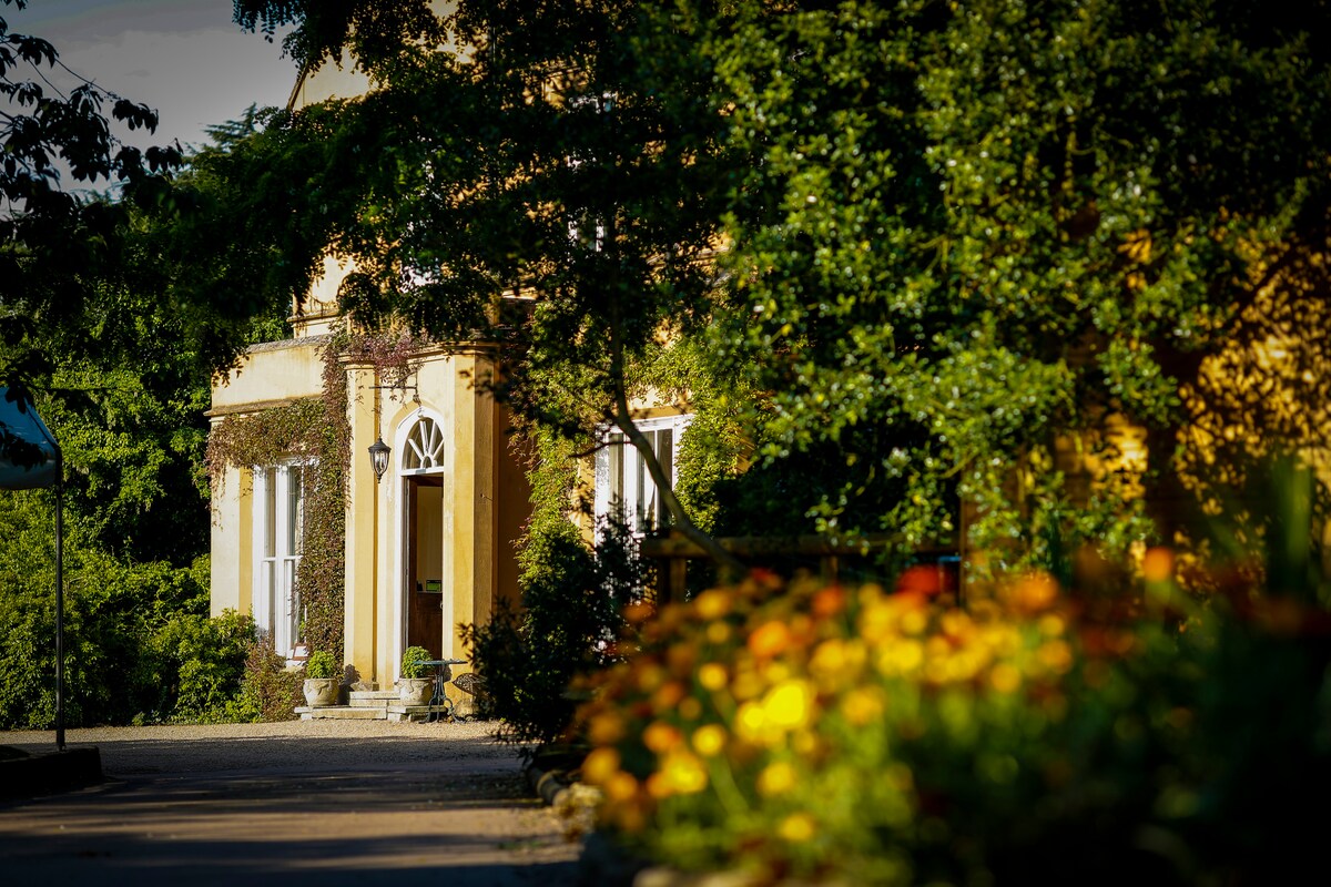 The entrance of a cottage is framed by lush greenery and colorful flowers. Symmetrical windows and a decorative door are visible, inviting guests into a warm and welcoming atmosphere. Soft sunlight filters through the trees, enhancing the charm of the surrounding garden.
