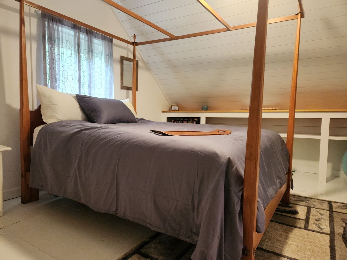 A four-poster bed is positioned in a softly lit room with sloped ceilings. The bed features a lavender comforter and two white pillows. A small mirror and a tidy workspace are visible in the background, with natural light filtering through a nearby window.