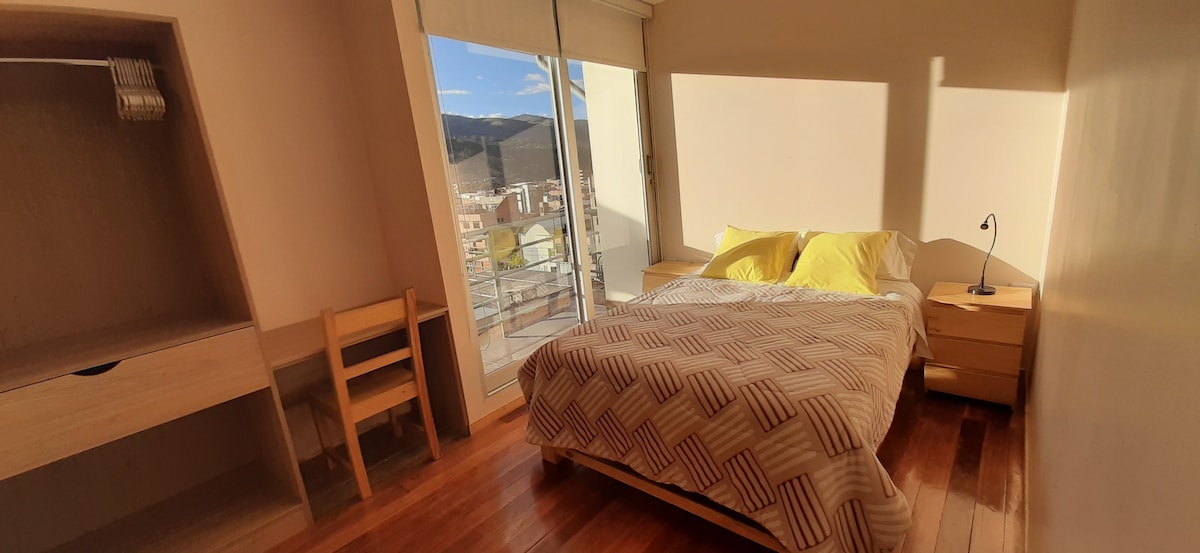 A well-lit bedroom features a spacious bed with yellow accent pillows. Two bedside tables and a desk with a chair are present, enhancing the functional layout. Large windows provide mountain views and allow natural light to fill the room.