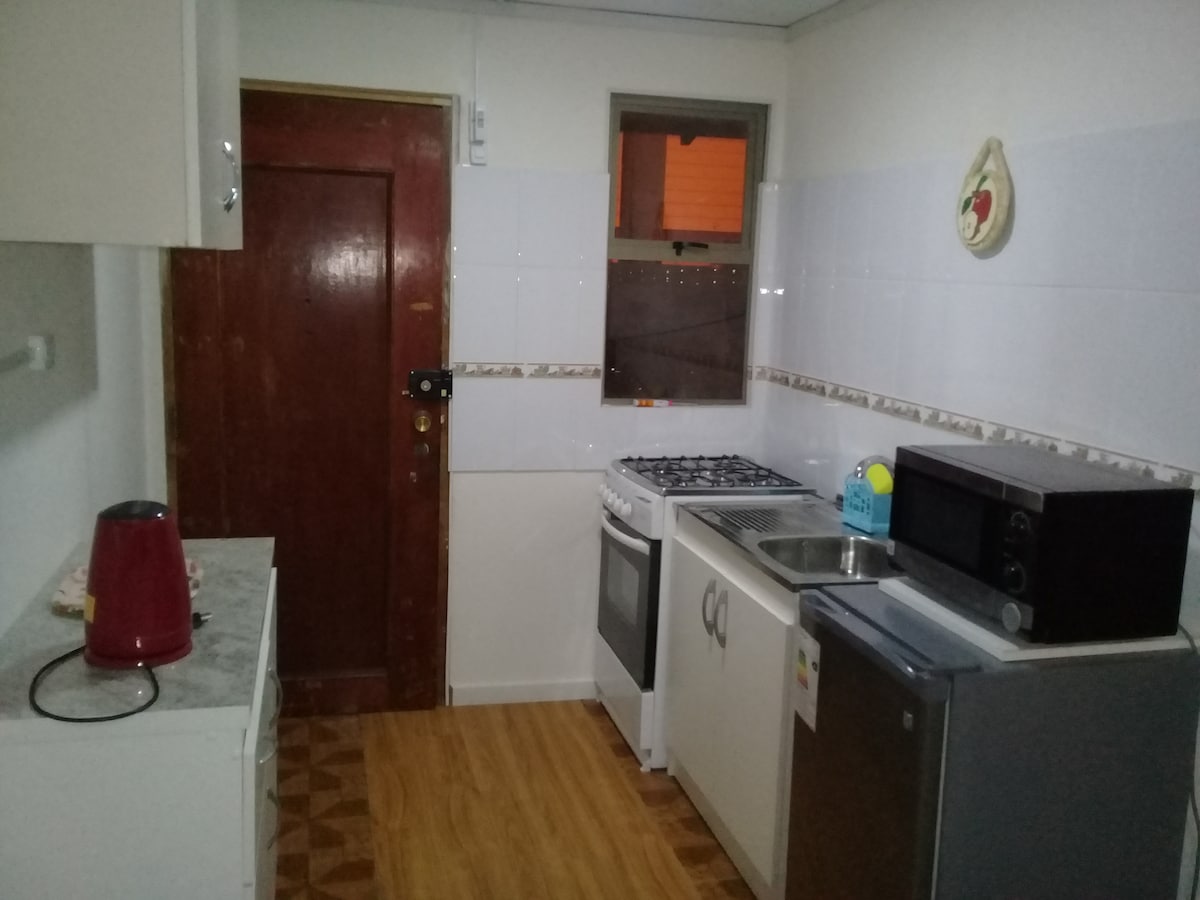 The kitchen features modern appliances including a stove and microwave, along with a refrigerator. A wooden door leads to the exterior, while cabinets and a countertop provide ample storage and preparation space. Light-colored walls and wooden flooring create a clean and functional environment.