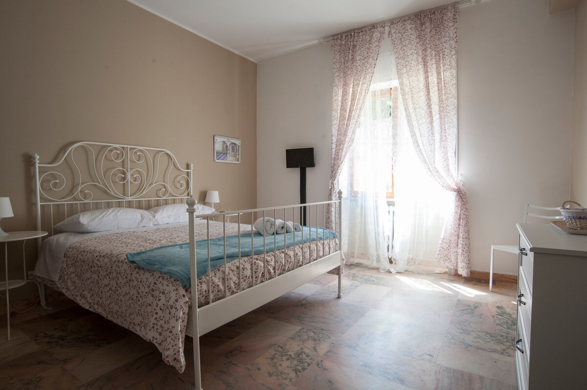 A sleeping area features a white metal bed with a floral patterned duvet and a soft blue throw draped over it. Natural light filters through sheer curtains, illuminating the room, while a dresser and bedside lamps complete the minimalist decor.