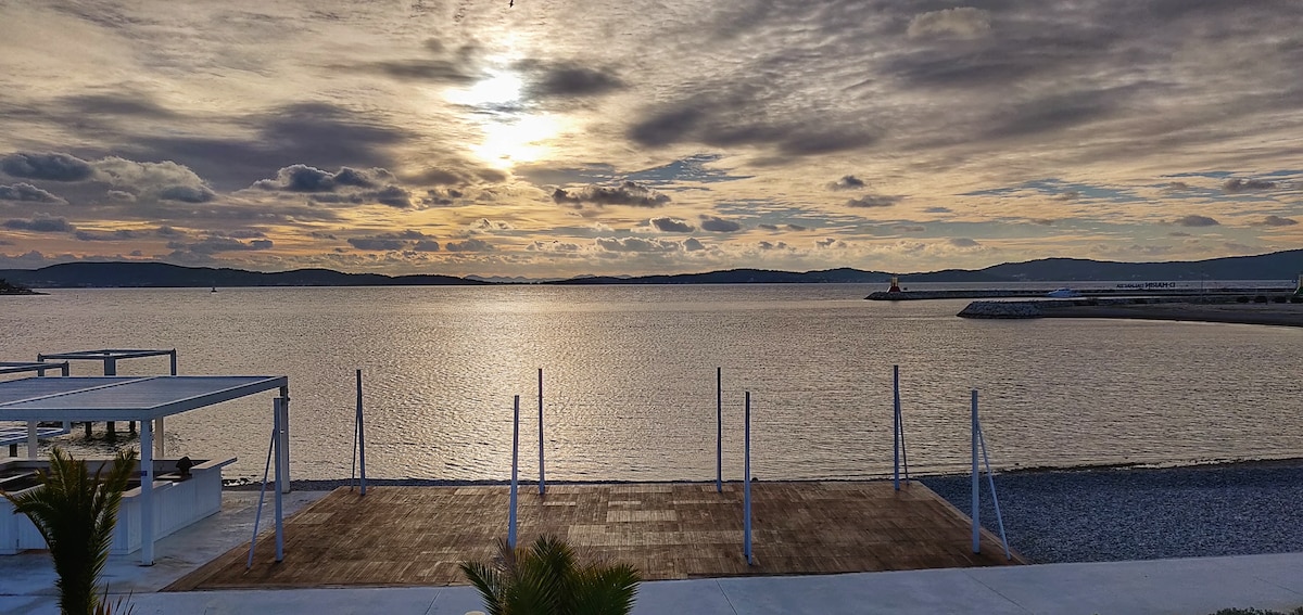 A wide view captures the tranquil waterfront, where the sun sets behind distant hills. Soft clouds reflect muted colors in the calm water, and wooden decking in the foreground connects to a pebbled beach. The scene conveys a sense of peaceful solitude.