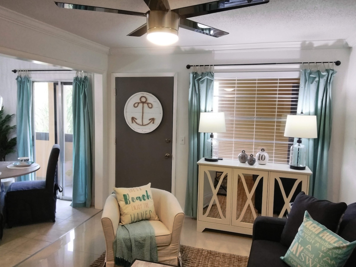 A living space features coastal-inspired decor, including a navy sofa and a light-colored armchair. Aqua curtains frame the windows, while a decorative anchor hangs on the wall. A dining table can be seen in the background, accompanied by a charming accent cabinet.