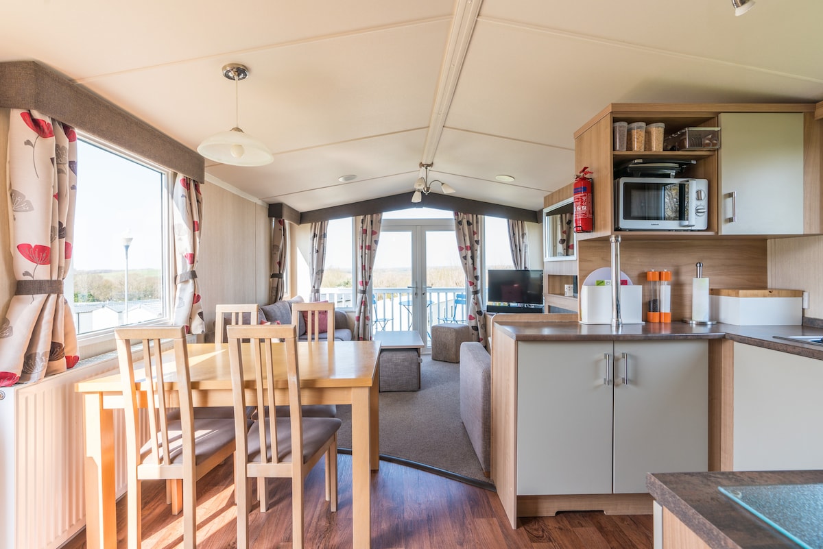 An open-plan living area is presented, featuring a dining table with six wooden chairs, next to a well-equipped kitchen that includes a microwave and other amenities. Large windows allow natural light to fill the space and offer views of the surrounding area.
