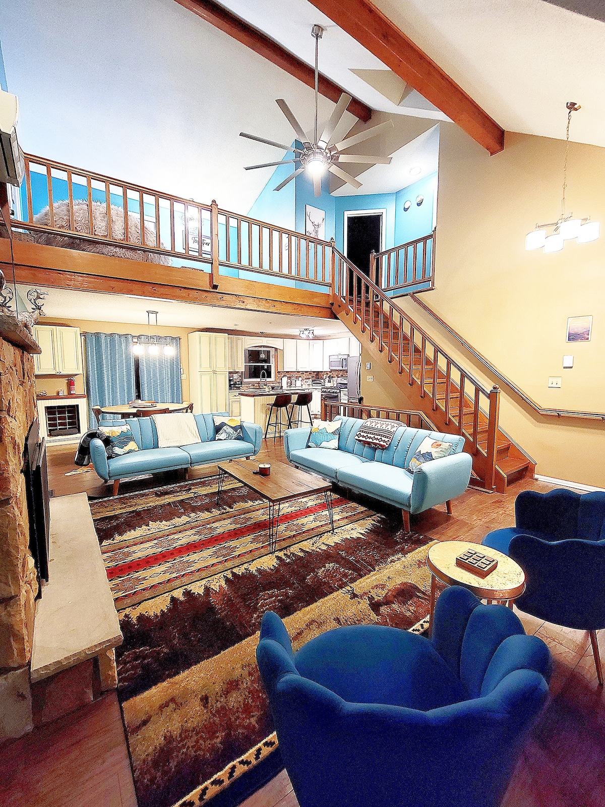 A spacious living area features high ceilings with exposed beams, accented by a large ceiling fan. Comfortable blue sofas and chairs are arranged on a patterned rug, offering a cozy seating arrangement. A staircase leads to the upper level, visible in the background.