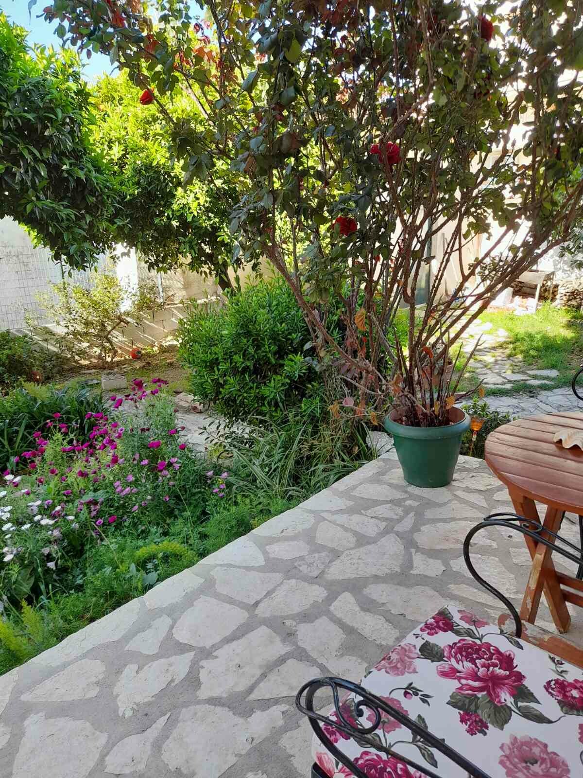 A stone patio area is framed by vibrant greenery and colorful flowerbeds. A table made of wood sits next to a decorative flower-patterned chair. Lush plants and blooming flowers, including pink and white petals, create an inviting garden space that enhances the outdoor setting.