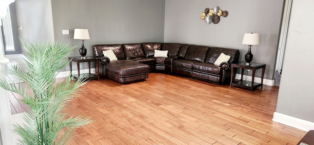 A spacious living area is furnished with a large dark leather sectional sofa, accompanied by two side tables with lamps. Light wood flooring enhances the bright atmosphere, while a decorative wall piece adds visual interest to the muted grey walls.