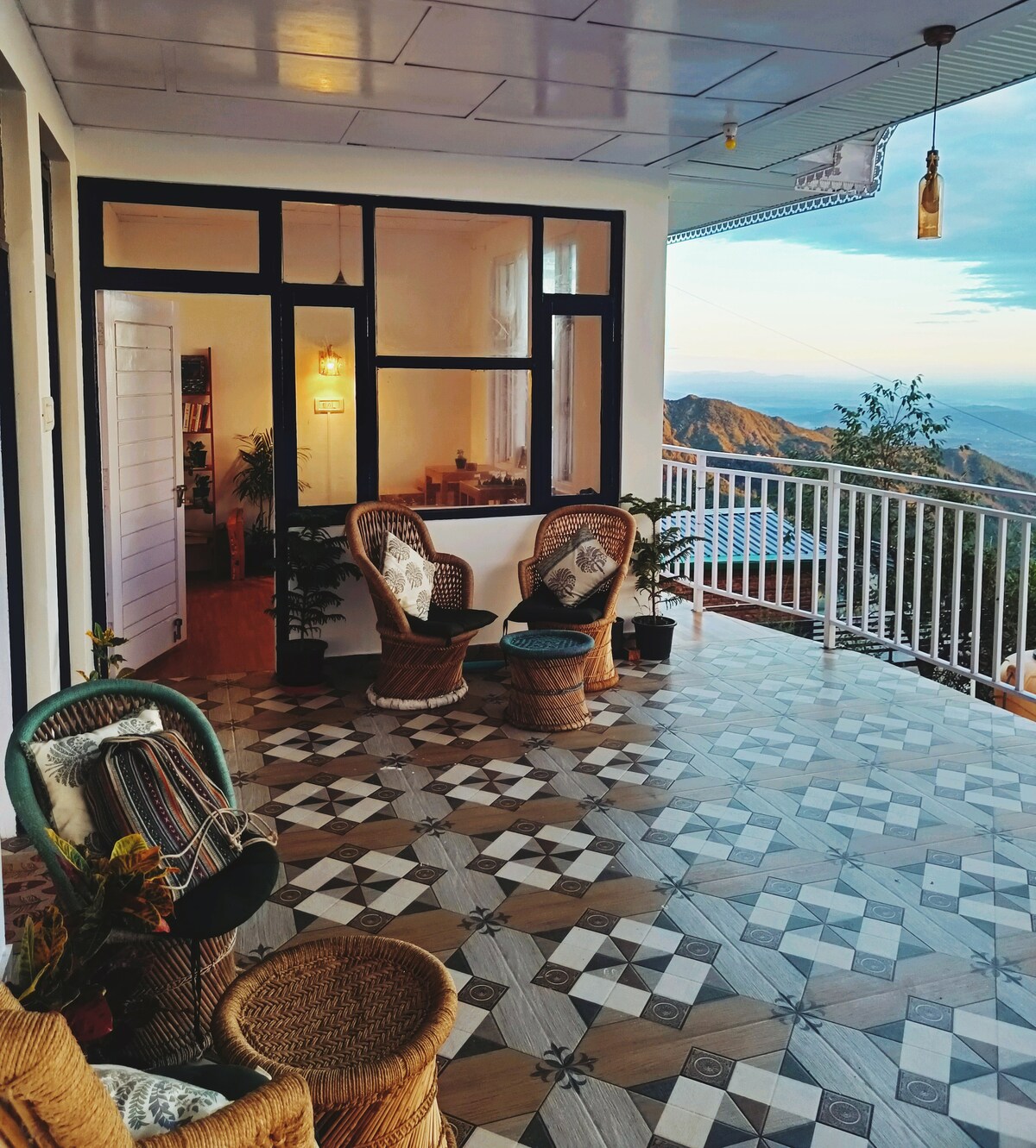 A spacious balcony is depicted, featuring two wicker chairs positioned on a patterned tiled floor. The area is filled with natural light, and potted plants are placed around. A gentle view of the surrounding hills can be seen in the background.