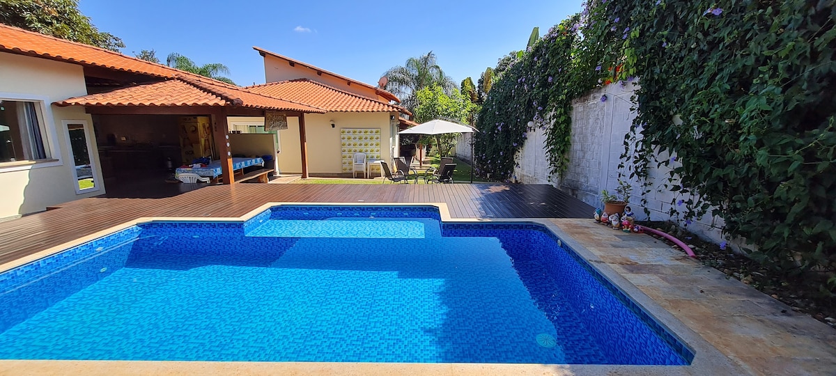 A inviting outdoor space features a clear blue pool set against a backdrop of lush greenery. A shaded area is provided by a spacious gazebo, with seating arranged nearby on a wooden deck. The house is visible, showcasing a tiled roof and large windows.