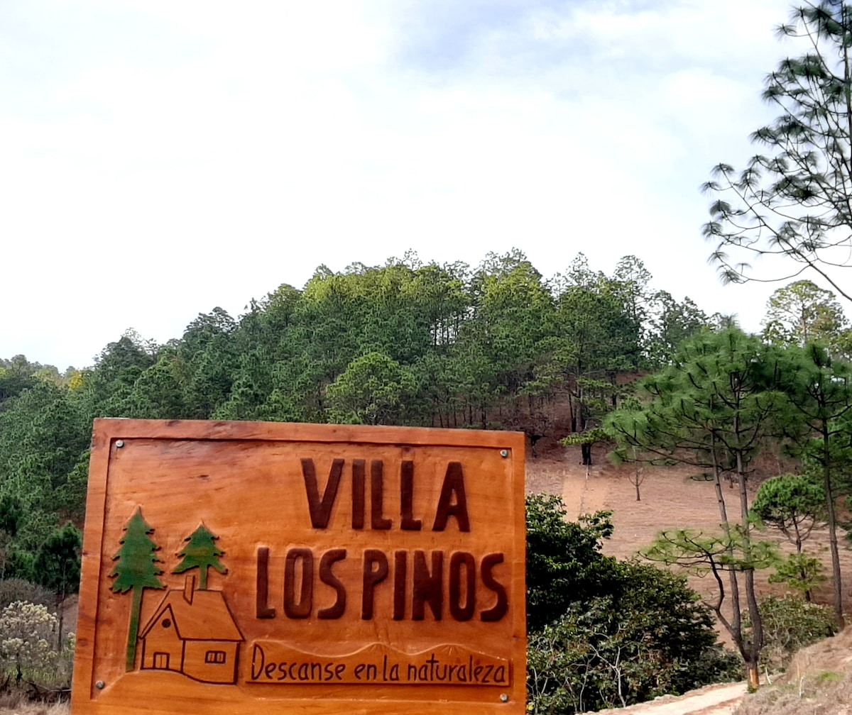 A wooden sign displaying 'Villa Los Pinos' is positioned prominently in front of a natural landscape. The background features a serene hillside with evergreen trees and varied vegetation under a partly cloudy sky.