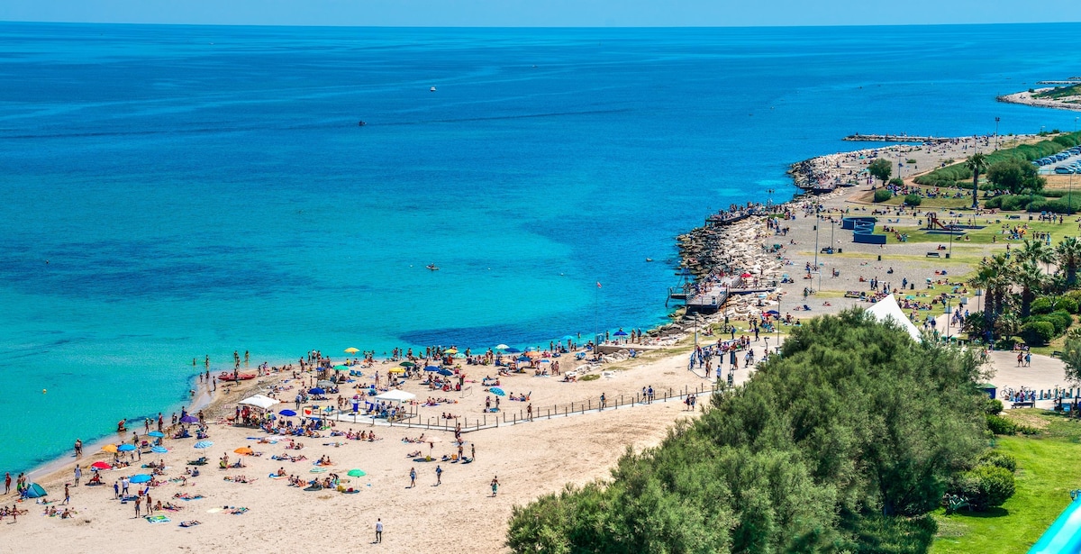 A panoramic view of a sandy beach is presented, featuring numerous sunbathers and umbrellas dotting the shoreline. The vibrant turquoise water gently meets the beach, with a rocky outcrop visible to the right. Lush greenery and recreational areas are seen in the background.