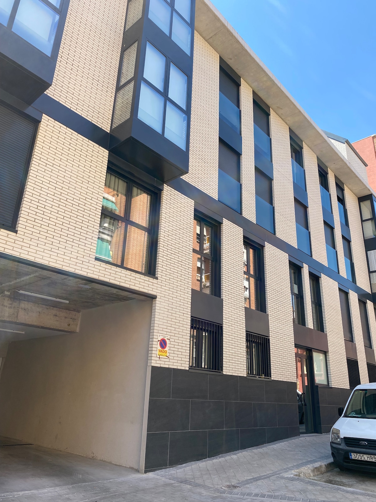 The modern building features a facade of light-colored brick and large windows, allowing ample natural light. The entrance is located under a covered parking area, indicated by a sign. The exterior design emphasizes a clean and contemporary aesthetic.