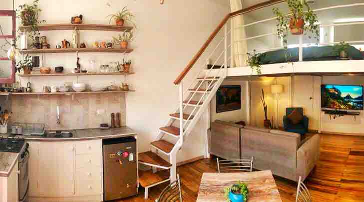 A modern kitchen area features open shelving with plants and various kitchen utensils. The dining space includes a table with chairs, while a cozy living area showcases a comfortable couch and a mounted TV. A wooden staircase leads to the bedroom area above.