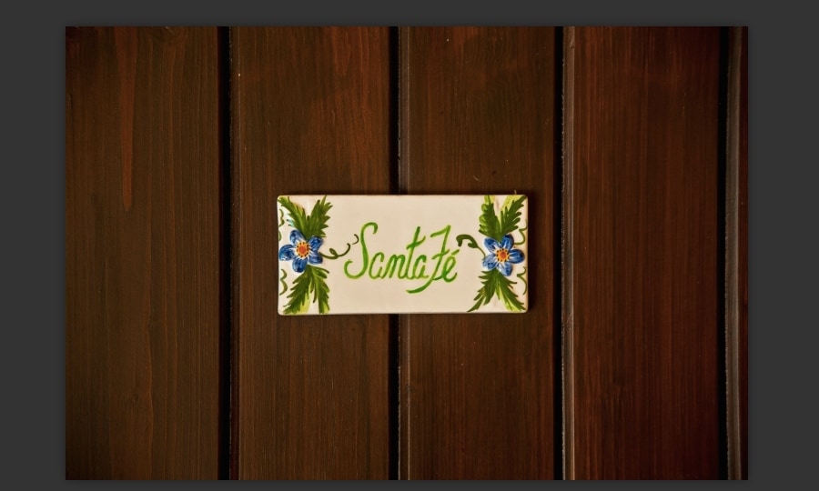 A decorative tile with the word 'Santa Fe' is mounted on a wooden panel, surrounded by floral motifs in blue and green. The rich brown tones of the wood create a warm backdrop, highlighting the elegant craftsmanship of the tile design.