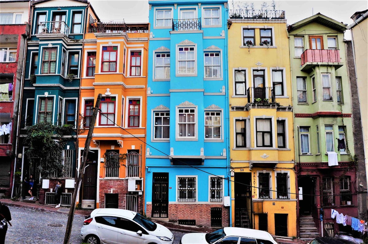 A row of colorful buildings lines a cobblestone street, showcasing vibrant hues of blue, yellow, and red. The structures feature ornate windows and balconies, with laundry hanging from some of them. A white car is parked along the curb, complementing the lively atmosphere.