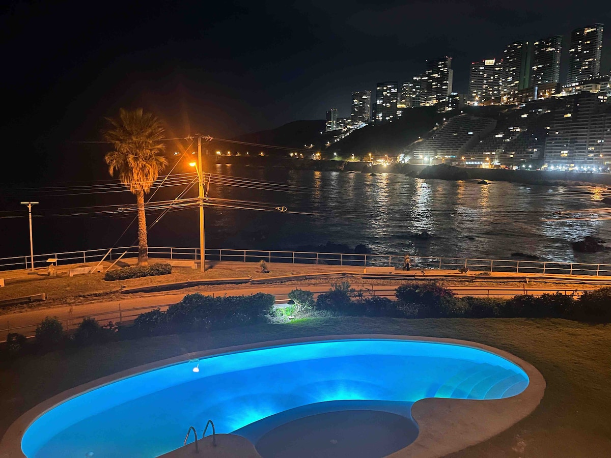 A serene outdoor pool glows softly in blue light at night, surrounded by lush greenery. The ocean shimmers in the background, with a distant view of illuminated skyscrapers along the shoreline, creating a calm and welcoming atmosphere by the coastline.