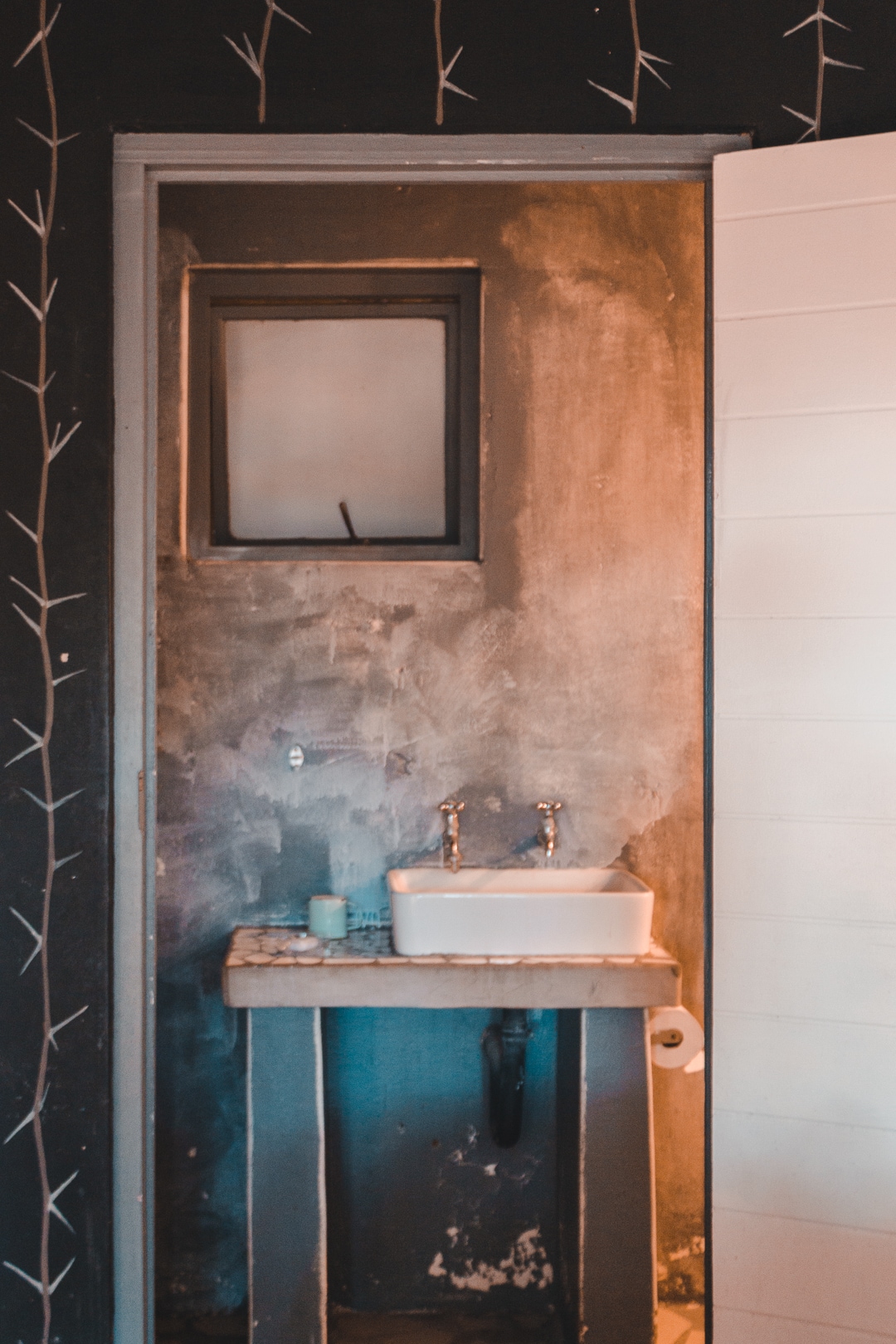 A simple bathroom setting is presented, featuring a small white basin mounted on a rustic countertop. Two metal faucets are visible above the basin. The walls are decorated with a subtle floral pattern, and a small window offers a glimpse of light into the space.