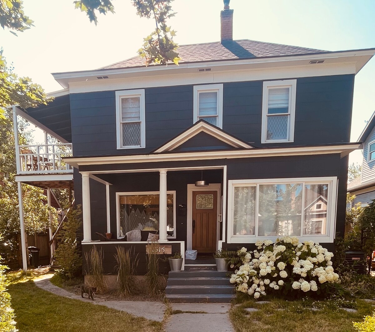 A charming two-story house features a dark exterior with white trim. The front porch is adorned with a welcoming wooden door, surrounded by large windows that invite natural light. Lush green plants and white flowering bushes enhance the front yard.