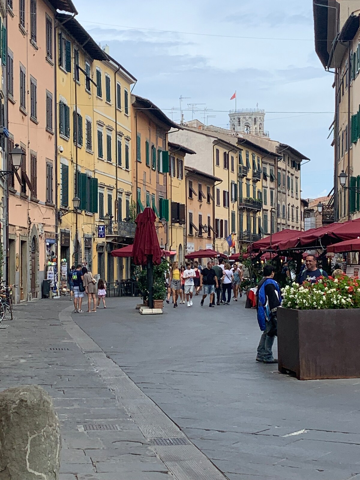 A lively street scene is showcased, featuring several pedestrians strolling past charming buildings with colorful facades and green shutters. Outdoor seating beneath red umbrellas invites visitors to enjoy the vibrant atmosphere. Flower boxes add a touch of greenery to the urban landscape.