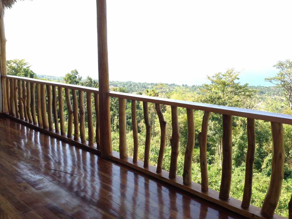 A spacious balcony features wooden railings with natural, textured posts. The polished wooden floor reflects sunlight, enhancing the serene jungle surroundings. An expansive view of the lush landscape and distant ocean is captured, providing a calm and quiet atmosphere.