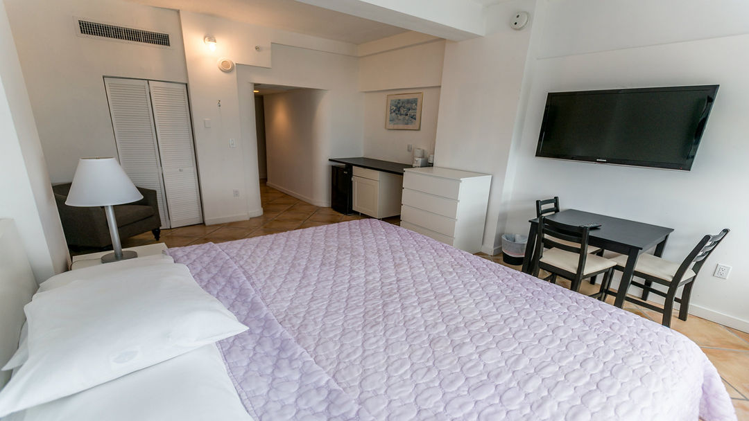 A spacious studio is highlighted by a large bed dressed in a lavender quilt. A kitchenette with a mini fridge is visible, alongside a dining area featuring a table and two chairs. A flat-screen TV is mounted on the wall.