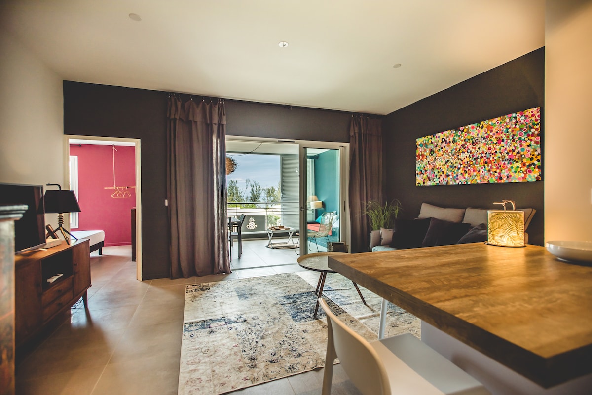 A modern living space is shown with a comfortable seating area adjacent to a dining table. Large sliding glass doors open to a balcony, offering views of the surroundings. Vibrant artwork decorates the wall, while soft curtains enhance the ambient lighting.
