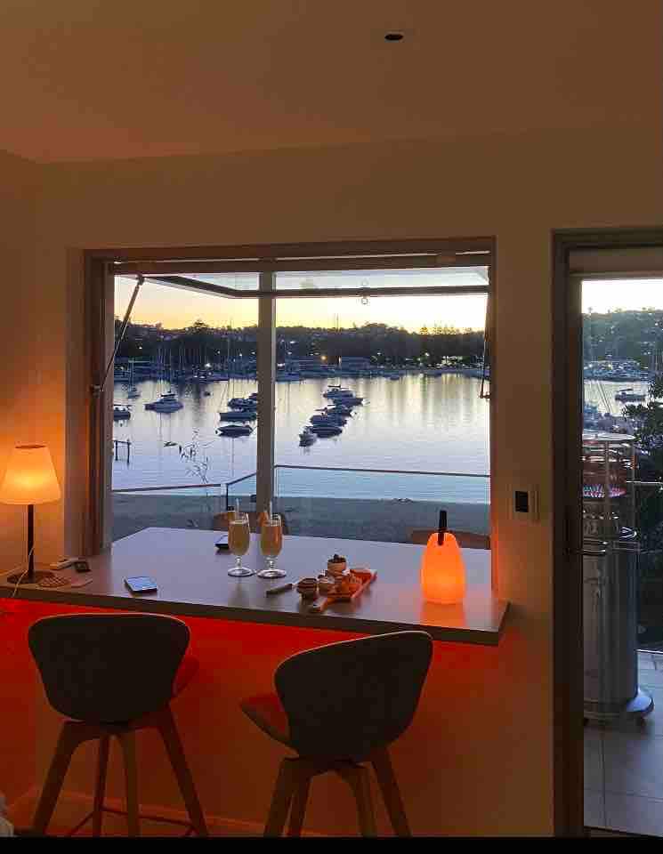 A window offers a scenic view of the marina, with boats gently bobbing in the water as the sun sets. A sleek bar counter, accompanied by two stools, is illuminated by soft lighting. A small assortment of snacks and beverages is positioned on the counter, adding to the ambiance.