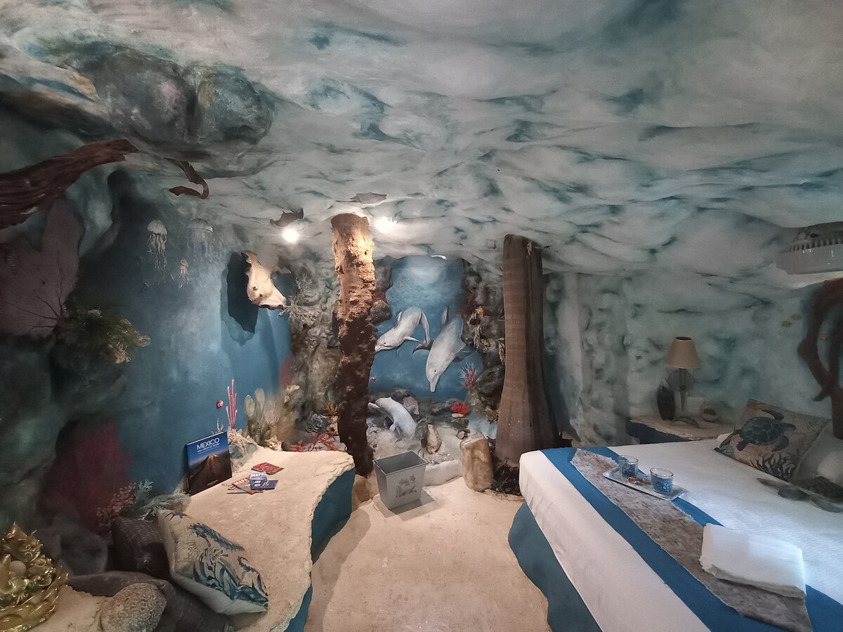 An ocean-themed room features walls and ceilings artfully painted to resemble an underwater environment. Natural elements like a tree trunk add character, while a spacious bed is positioned alongside decorative pillows. Soft lighting enhances the tranquil atmosphere, creating a serene retreat.