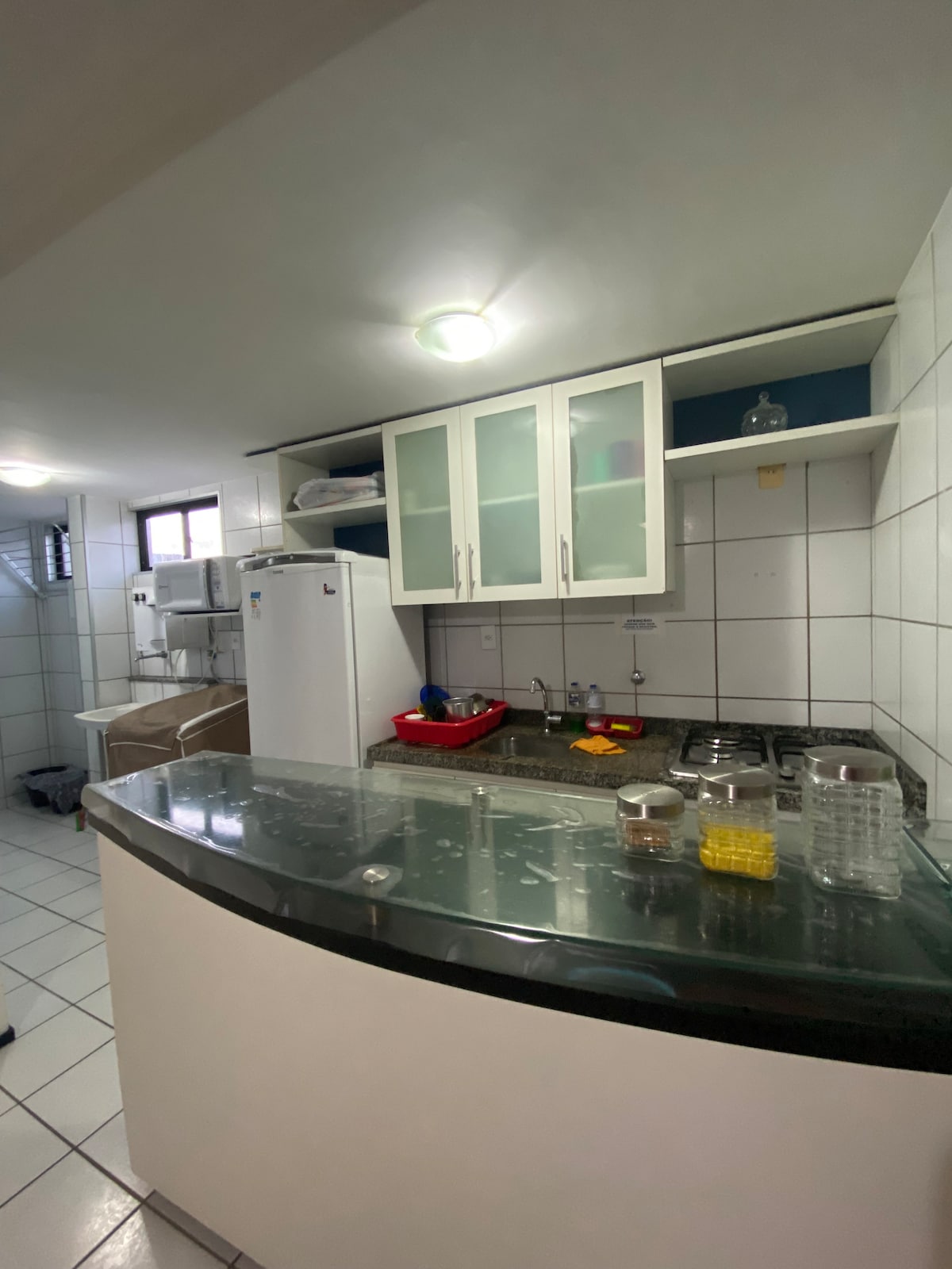 The kitchen features a curved, dark countertop with a white base and cabinetry above. A refrigerator and a stove are visible, alongside a red tray with kitchen essentials. Shelves display various items, and tiled walls contribute to a clean, organized environment.