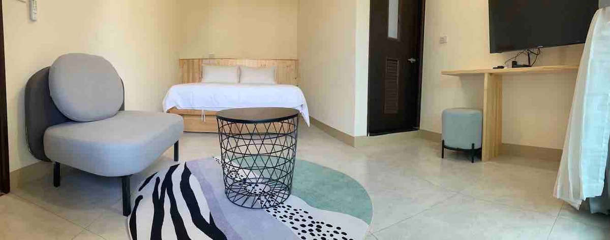 A light-filled room features a spacious bed with white linens and a cozy seating area with a soft, rounded chair. A circular wire coffee table sits on a textured area rug, while a wall-mounted television is positioned for easy viewing.