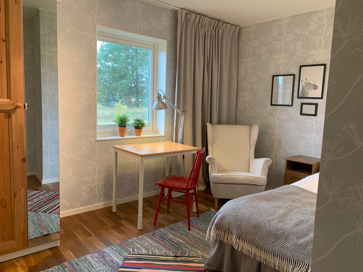 A bright room features a spacious bed draped with a soft blanket and a cozy armchair. A desk and a red chair are positioned by the window, which offers a view of greenery outside. The walls are adorned with subtle botanical wallpaper.