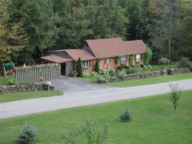 A charming bungalow is portrayed amidst lush greenery, featuring a sloped roof and a well-maintained garden. A gravel driveway leads to the entrance, framed by a stone wall. Surrounding trees add to the serene, natural setting.