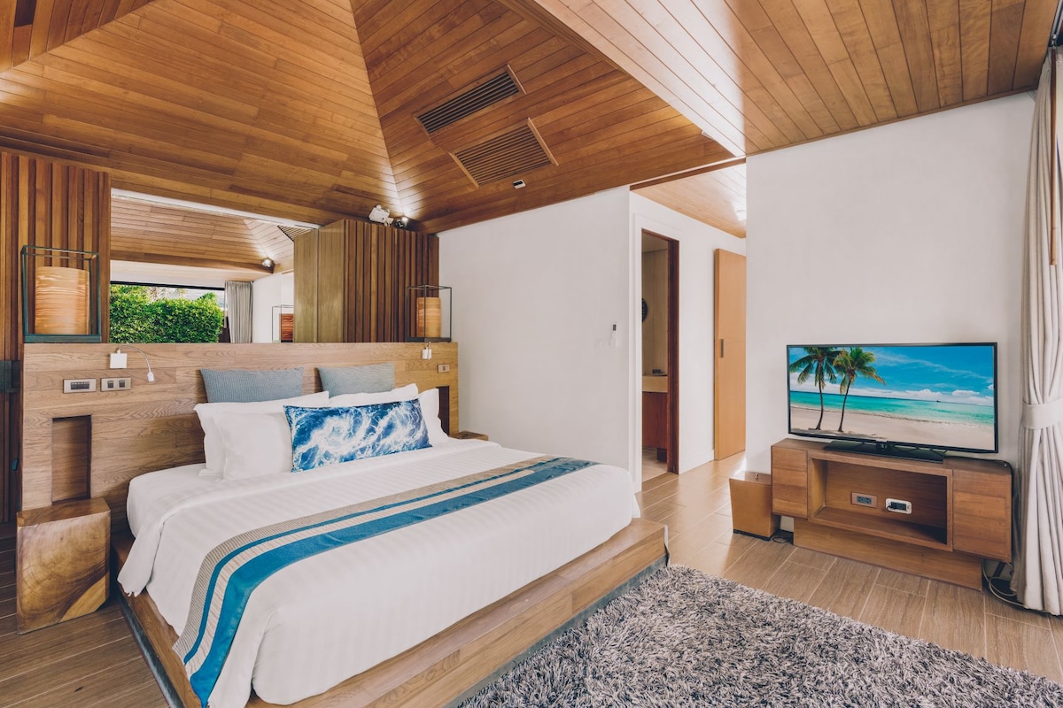 A spacious bedroom is featured, showcasing a large bed with crisp white linens and decorative pillows. Natural wood elements enhance the design, complemented by a plush area rug. A flat-screen television is positioned on a low wooden console, with ambient light cascading through nearby windows.