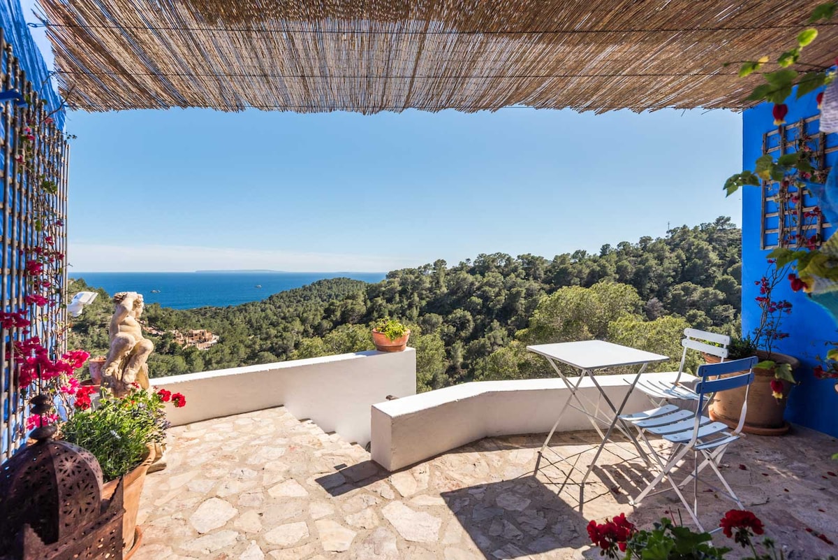 An outdoor terrace features a stone floor adorned with potted plants and flowers. A small white table and two blue chairs are arranged for seating, with sweeping views of the mountains and sea visible in the background under a clear blue sky.