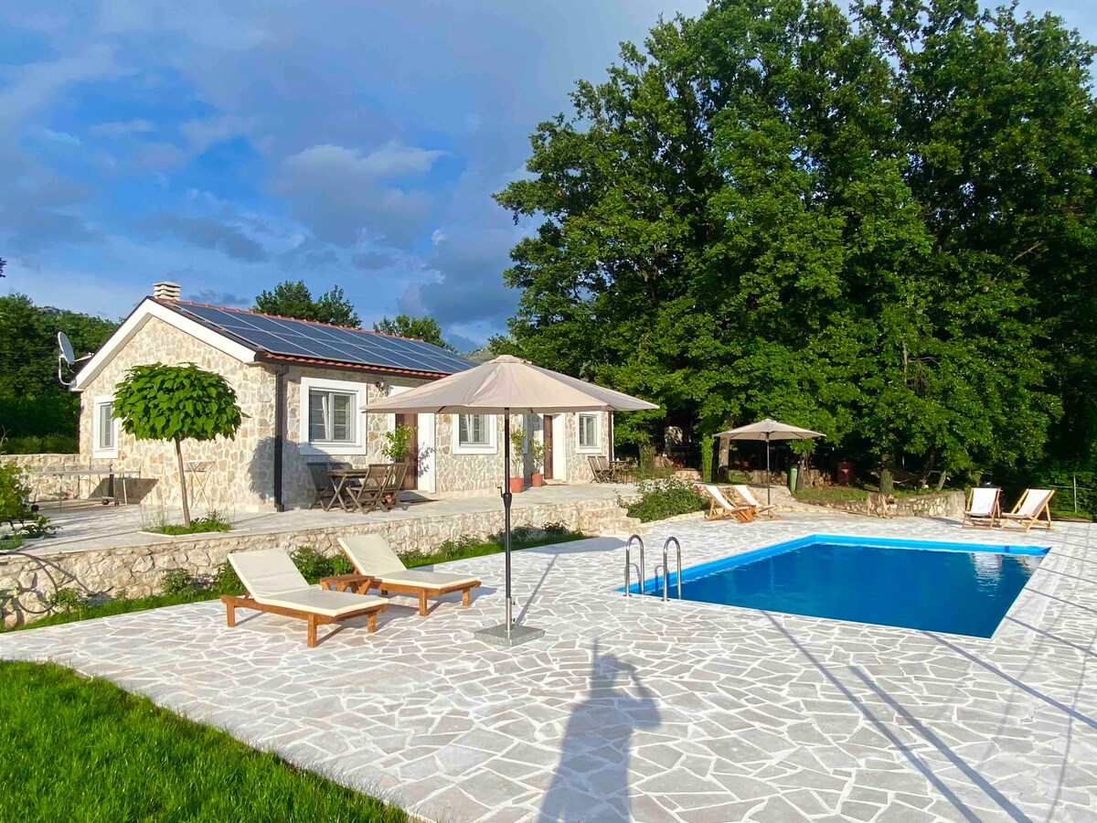A charming stone villa with large windows is surrounded by lush greenery. A refreshing pool sits nearby, accompanied by sun loungers and an umbrella. The patio area features a table and chairs, creating a welcoming spot for outdoor meals.
