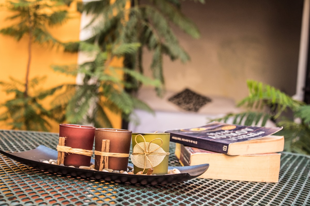 A decorative tray holds three candles wrapped in natural materials, placed alongside a stack of books. In the background, green plants provide a refreshing touch, enhancing the outdoor area’s peaceful ambiance.